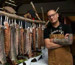 Chef Shane Chartrand in an apron beside smoked fish hanging to dry